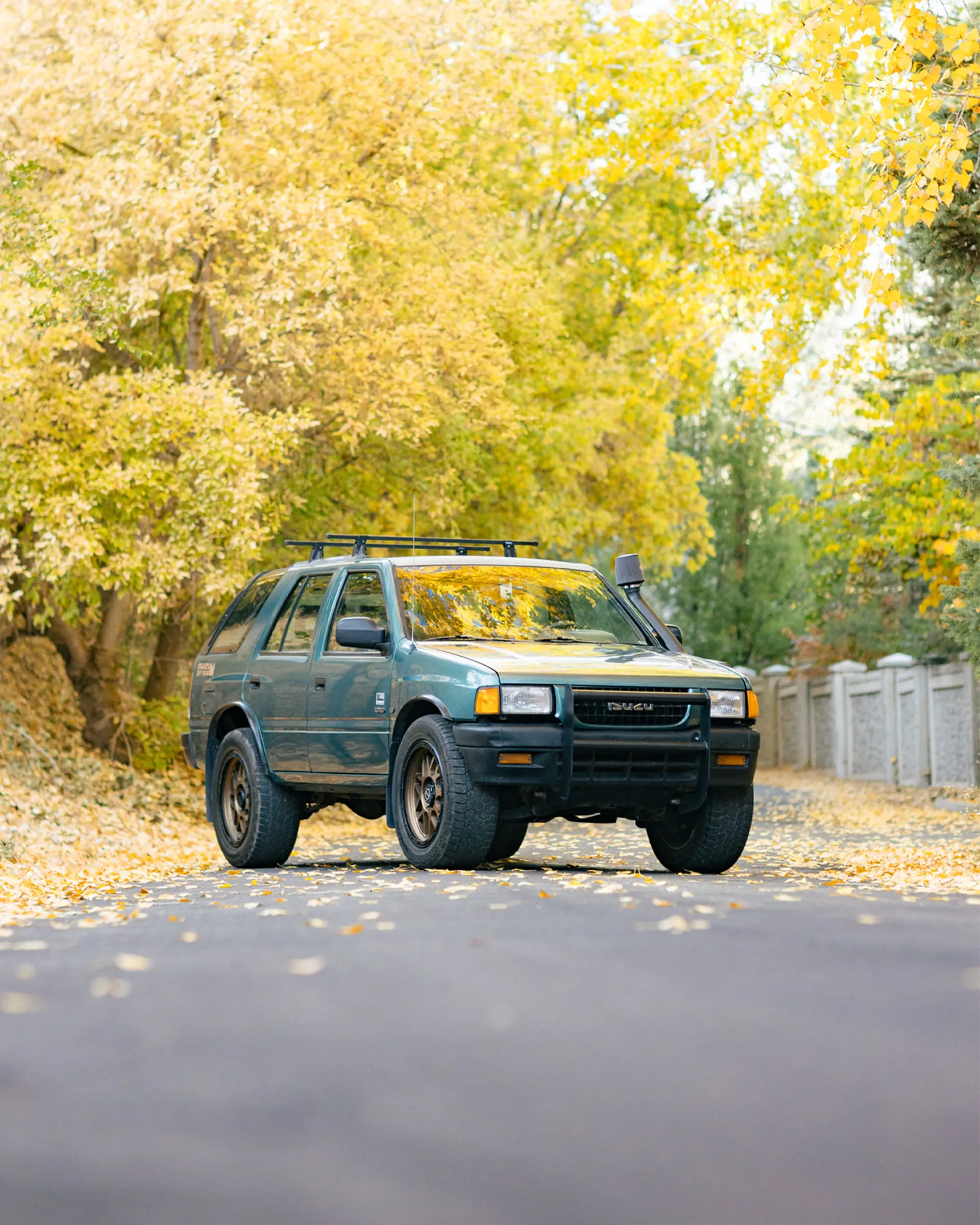 Green Isuzu Rodeo parked on a leaf-covered lane under autumn trees